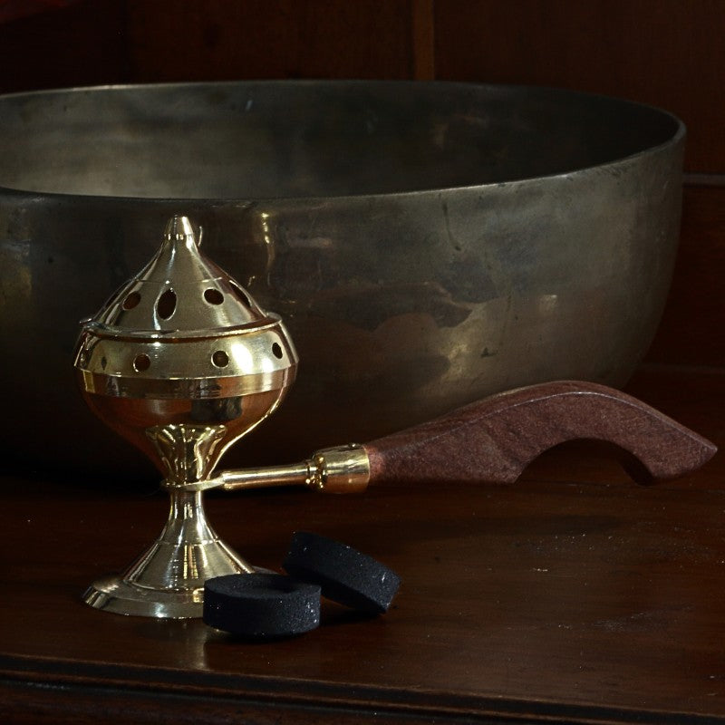 Brass charcoal incense burner with lid and wooden handle next to a brass singing bowl and charcoal discs