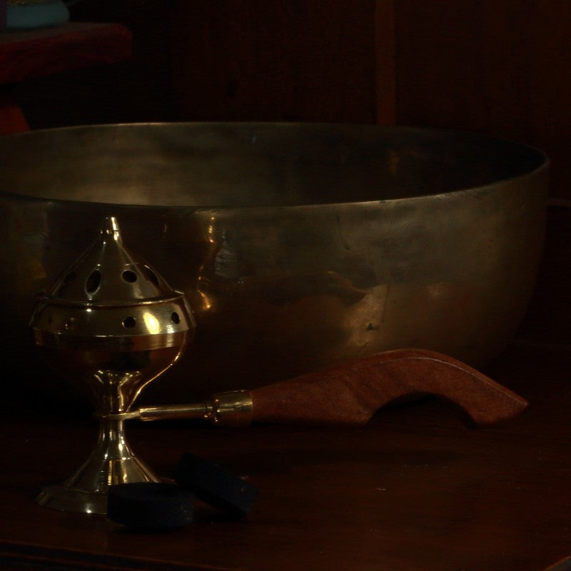 Brass charcoal incense burner with lid and wooden handle next to a brass singing bowl and charcoal discs
