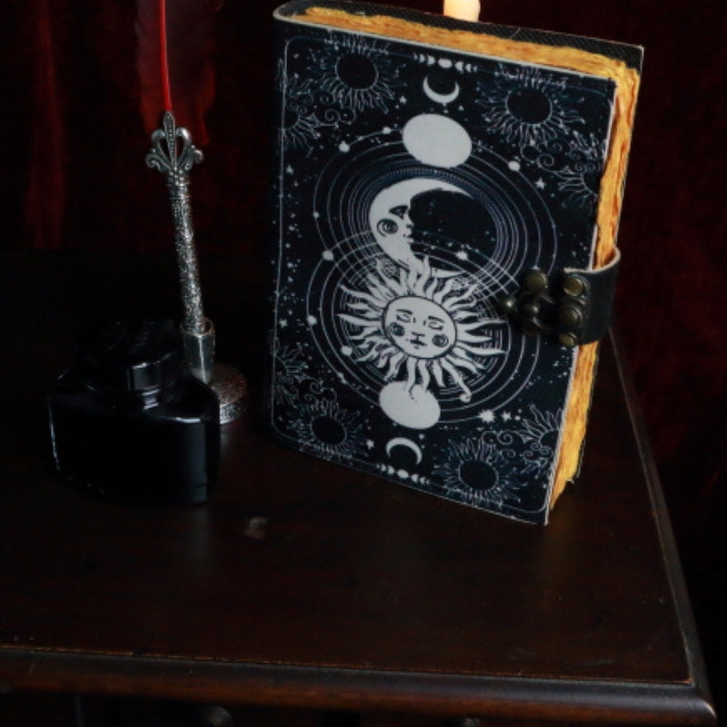 leather bound book with an image of a sun and moon on the front. A red feather pen and ink sitting next to the book,