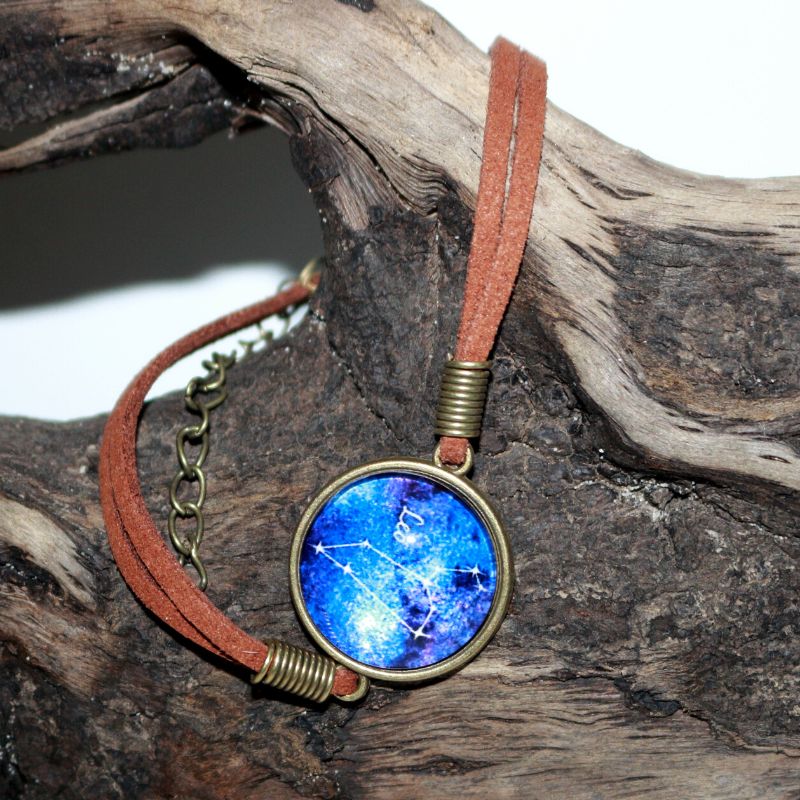 leather bracelet with a blue glass cabochon bead in the centre with the line drawing of the constellation of leo and the word leo written in white. The bracelet hangs on a piece of driftwood.