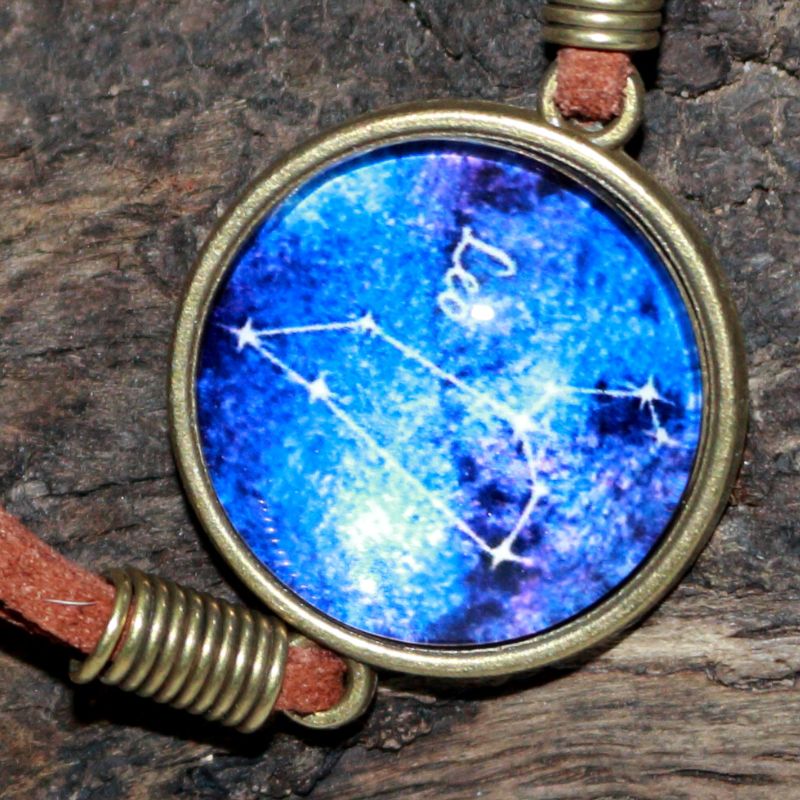 leather bracelet with a blue glass cabochon bead in the centre with the line drawing of the constellation of leo and the word leo written in white. The bracelet hangs on a piece of driftwood.