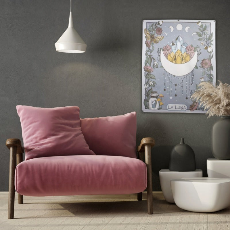 wall hanging of a crescent moon with points facing upwards, crystals and flowers inside the space between the points. The rectangular tapestry is bordered with vines, flowers and crystals. Tapestry is hanging on a grey wall behind a wooden chair with pink cushions. in front of the tapestry is 3 white abstract tables, 2 contain dark grey vases and 1 has brown grass in it. There is a white light hanging from the ceiling