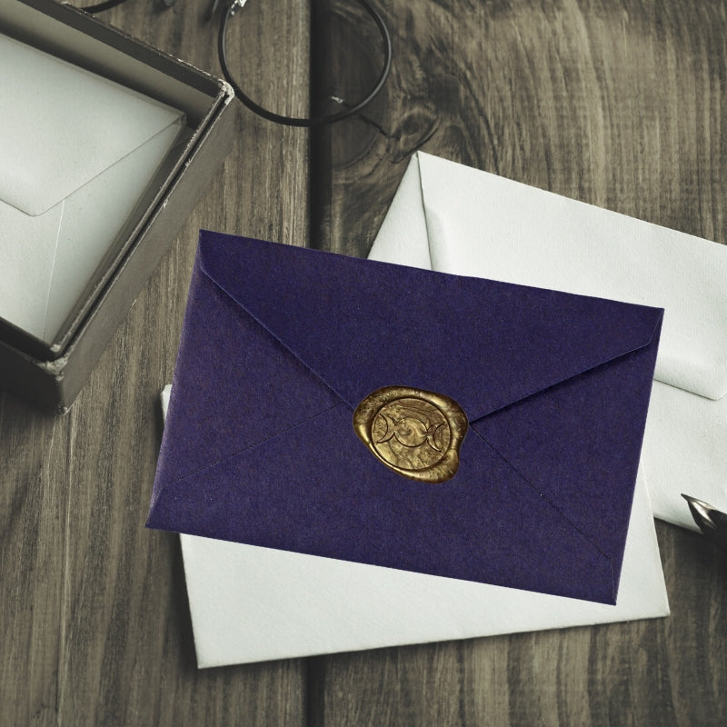gold triple moon wax seal on a purple envelope, sitting on a wooden table with white envelopes
