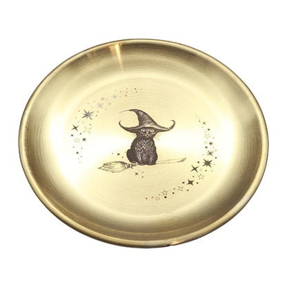 A round, shallow gold-colored metal dish featuring a central illustration of a black cat wearing a wizard hat and sitting on a broomstick, surrounded by small stars.