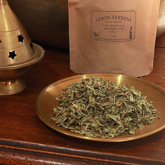 A kraft paper pouch labeled 'LEMON VERBENA' stands behind a brass plate filled with dried green herb leaves on a wooden surface.