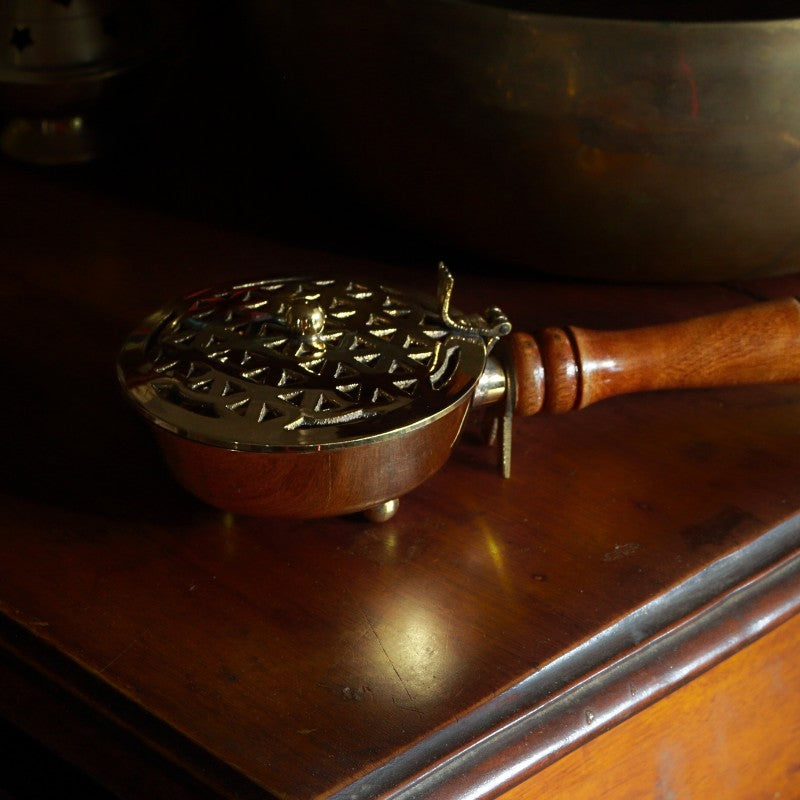 Brass charcoal burner with wooden handle sitting ion a wooden table in front of a brass singing bowl