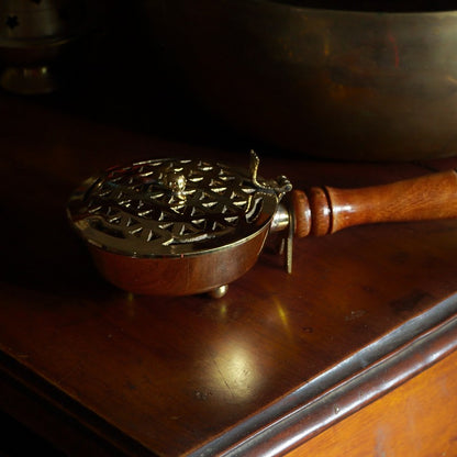 Brass charcoal burner with wooden handle sitting ion a wooden table in front of a brass singing bowl