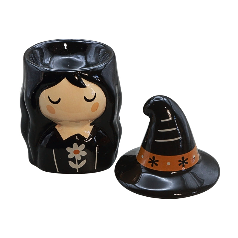 A black witch-themed ceramic candle holder set with a body shaped like a sleeping witch's head with long black hair and a flower motif, alongside a separate pointed witch hat lid with an orange band and black star details, on a white background.