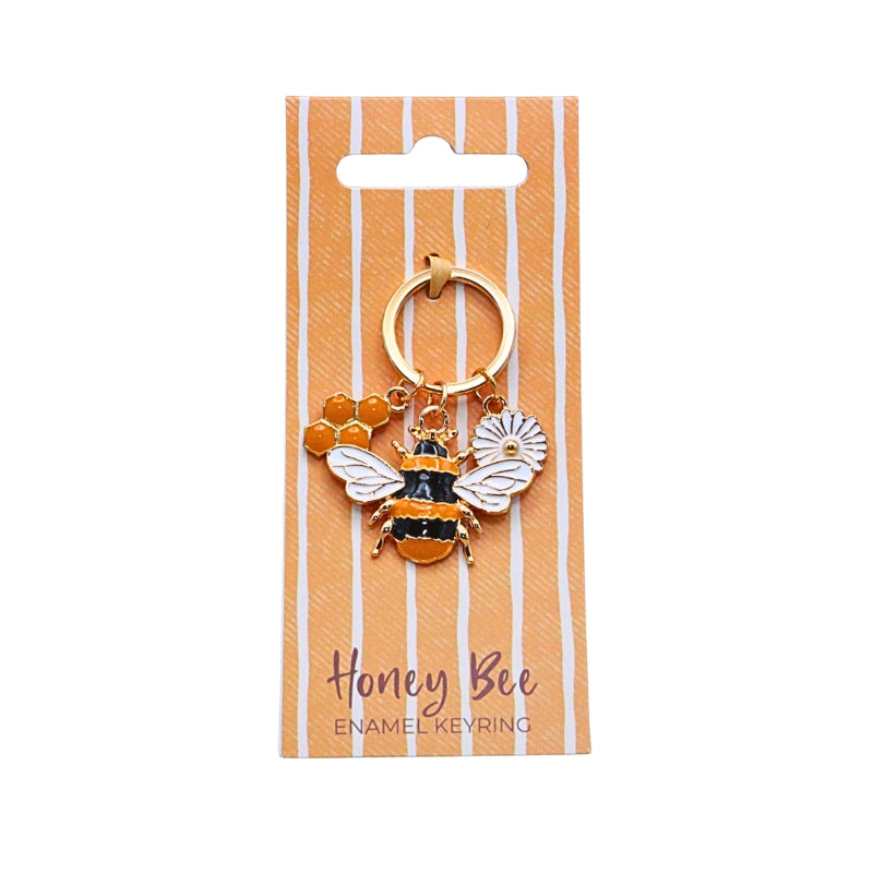 A gold-tone enamel keyring featuring a honey bee, a flower, and a honeycomb charm, displayed on a peach and white striped card labeled 'Honey Bee Enamel Keyring'.
