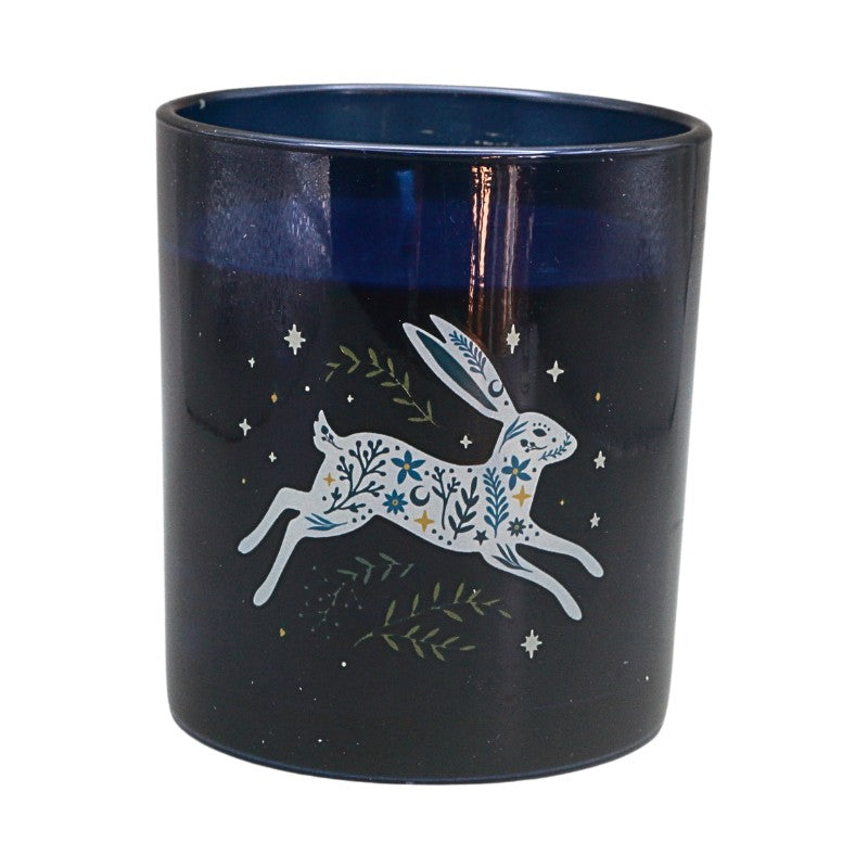 A dark navy blue glass candle jar featuring a white line-art illustration of a hopping rabbit decorated with blue and green floral patterns, surrounded by small stars and leaves.