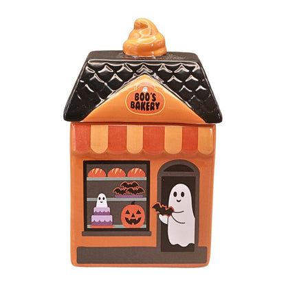 A Halloween-themed ceramic cookie jar shaped like 'Boo's Bakery'. The jar features an orange facade, a black textured roof with a small orange pumpkin topper, a striped awning, a window displaying a ghost, pumpkin, and cake, and a door with a white ghost handing out cookies.