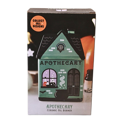 A boxed ceramic oil burner shaped like a haunted house labeled 'APOTHECARY,' shown on a plain white background. The front of the box features a teal house illustration with black roof and door details, an orange circular badge reading 'COLLECT ALL DESIGNS,' and the text 'APOTHECARY CERAMIC OIL BURNER' near the bottom.