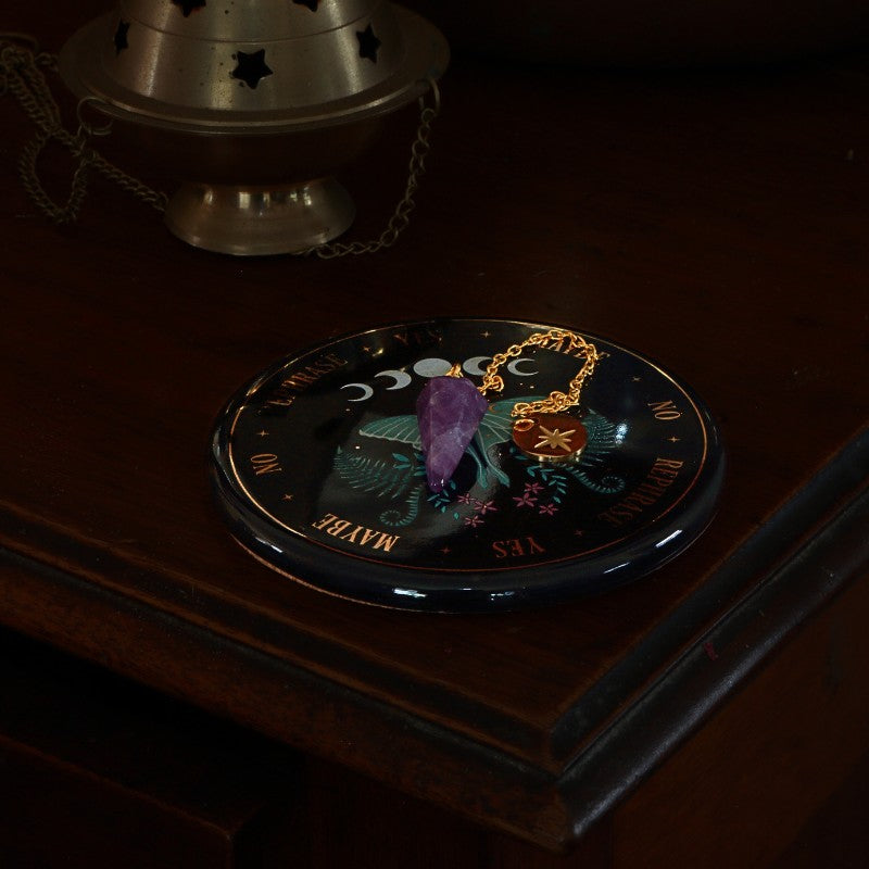 Decorative plate with celestial design  and amethyst pendulum 
on a dark surface