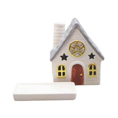 A small decorative house-shaped object with a gray roof, white body, red door, gold window trim, black stars, and a pentagram emblem on the front, shown alongside a rectangular white tray on a plain white background.