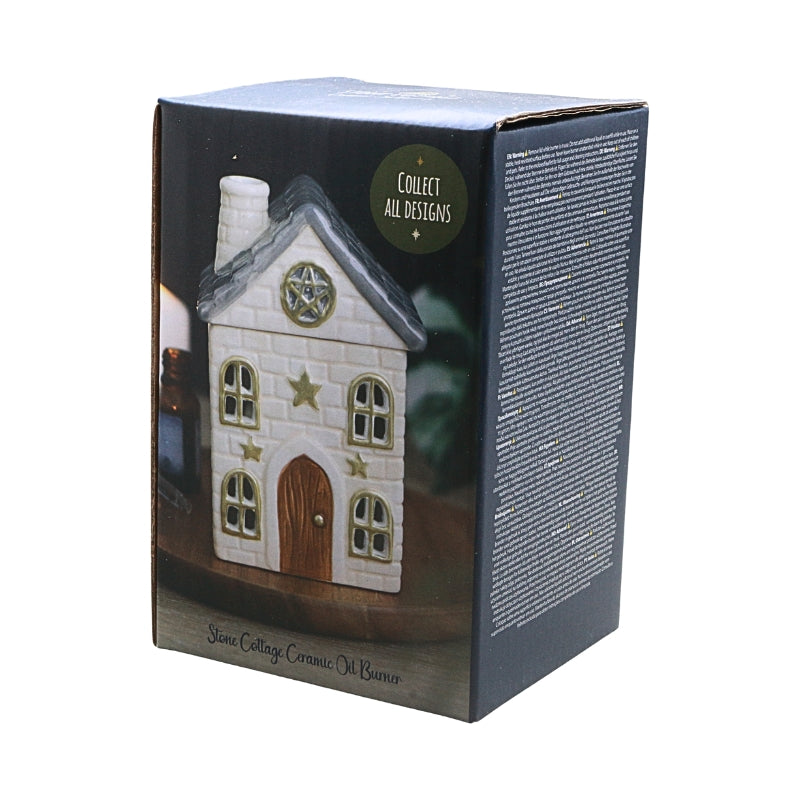 A boxed collectible ceramic oil burner shaped like a small white cottage with a gray roof, gold stars, and a brown door, shown on a plain white background. The front of the box features a photo of the burner and a green circular badge reading 'Collect All Designs.