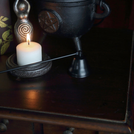 apothecary cabinet with a spellcrafting book on top, a black cast iron cauldron with a pentcle design on front, a black candle snuffer, candle holder in the shape of a goddess holding a triple moon.