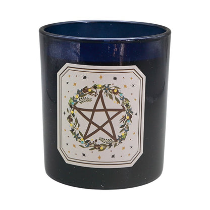 A dark navy blue glass jar candle with a white decorative label featuring a brown pentagram inside a floral wreath, set against a plain white background.