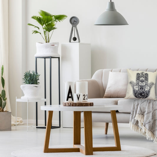 modern lounge room with white furniture and wood and black accents- a white lsofa with a grey throw rug, white cushion and linen white cushion with a black hamsa symbol printed on front of it. Green plants in background